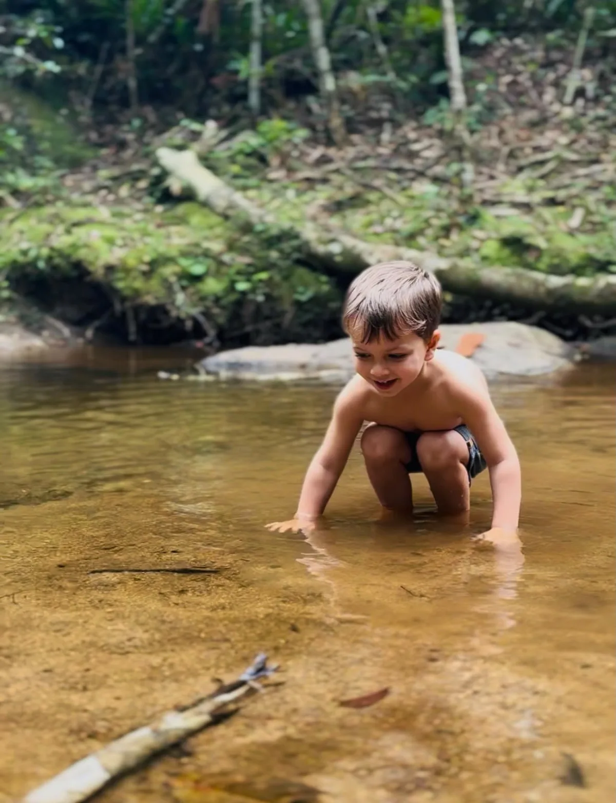 Criança agachada dentro de um trecho raso de água transparente, com mata ao fundo.