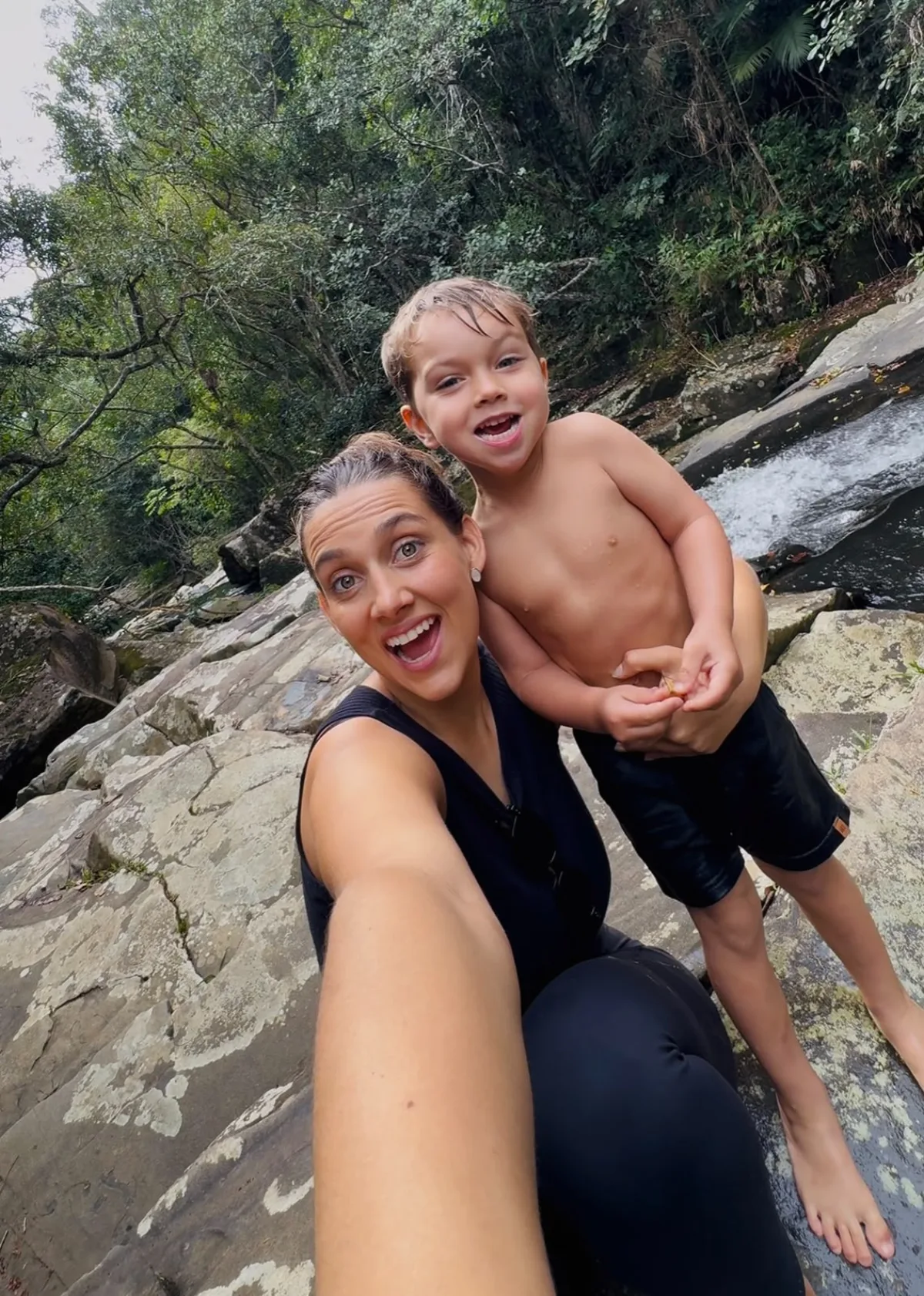 Miriá e uma criança sentados juntos na laje de pedra ao lado da corredeira.