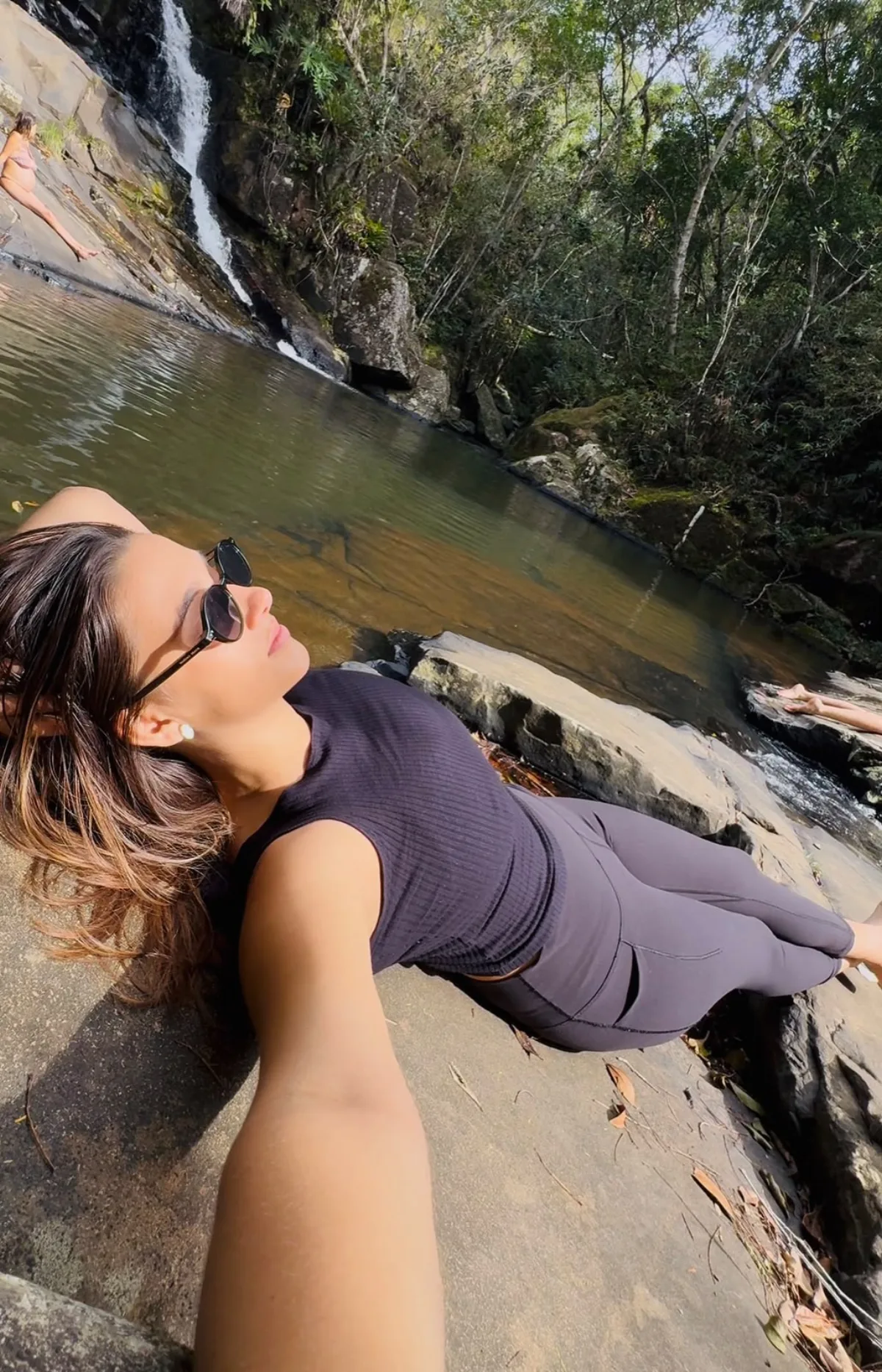 Miriá deitada na pedra à beira do poço, com a queda d’água ao fundo.
