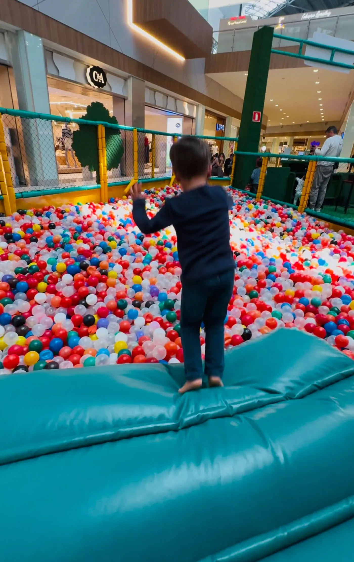 Criança pequena de costas, em pé sobre uma borda inflável, diante de uma piscina de bolinhas coloridas dentro de um shopping.