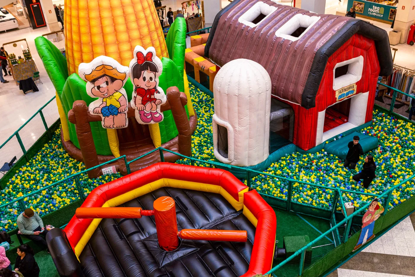 Vista de cima do brinquedo inteiro: piscina de bolinhas, celeiro, silo e uma arena inflável cercados no vão do shopping.
